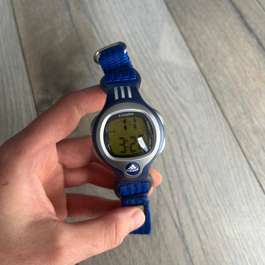Adidas 2000s Blue 10-0117 Digital Watch (model X-003) is a blue digital watch with a silver bezel and ergonomic case. It features a blue nylon Velcro strap, four side buttons, and a blue-green backlight display showing Adidas logo. The design includes three white stripes on the case and a silver Adidas logo button below the screen. The stainless steel back is engraved and marked water resistant up to 3 ATM, with an Adidas logo patch on the strap.