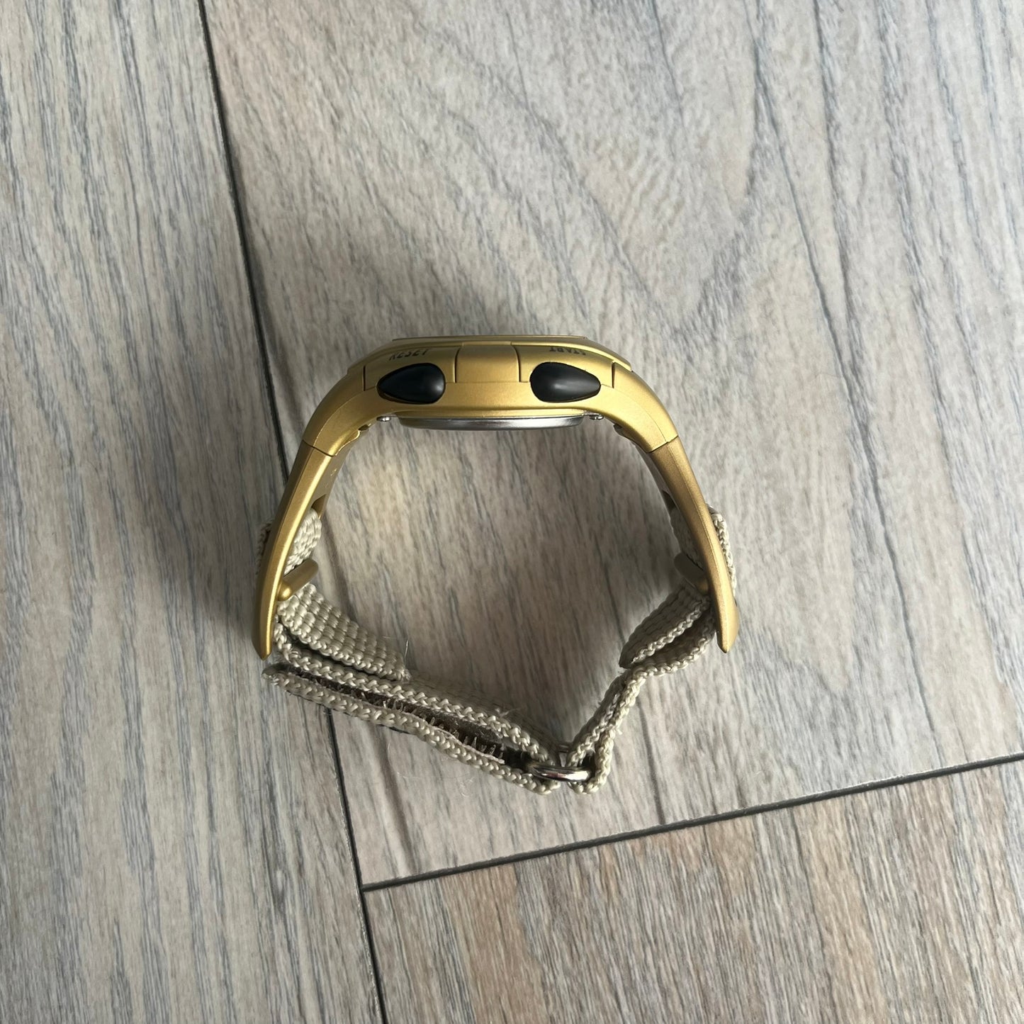 Adidas 2000s Gold 10-0117 Digital Watch (model X-003) is a fully gold digital watch with big and ergonomic case. It features a gold plastic and nylon strap, four side buttons, and a blue-green backlight display showing Adidas logo. The design includes three black stripes on the case and a black Adidas logo button above the screen. The stainless steel back is engraved and marked water resistant up to 3 ATM.