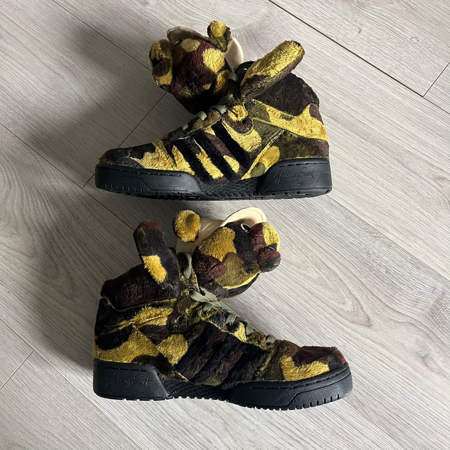 Adidas Jeremy Scott “Camo Bear” Sneakers (Style Q20917) released on March 16, 2013, in Earth Green/Black/Shioli. Featuring plush fleece uppers with a camouflage pattern, black three stripes, and a fully stuffed teddy bear head and arms on the tongue. Includes Jeremy Scott signature detailing on the inside heel and a black rubber sole.