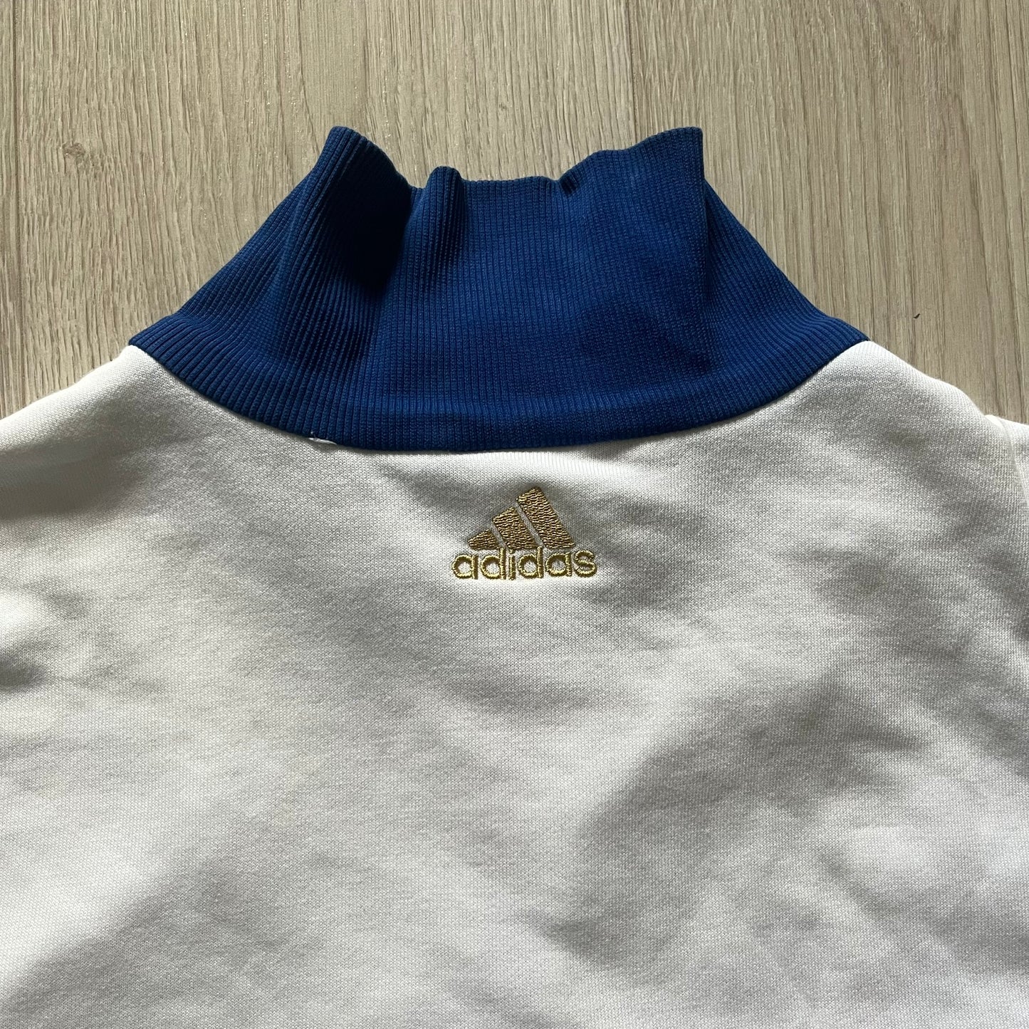 Adidas USA Olympic London 2012 Track Jacket in white with a blue sleeve featuring bronze, silver, and gold adidas stripes. Designed with a red zipper, blue collar, and embroidered adidas USA logo in a flag graphic on the chest, paired with a gold embroidered London 2012 logo on the other side. A rare piece of official Team USA gear from the London Olympic Games.