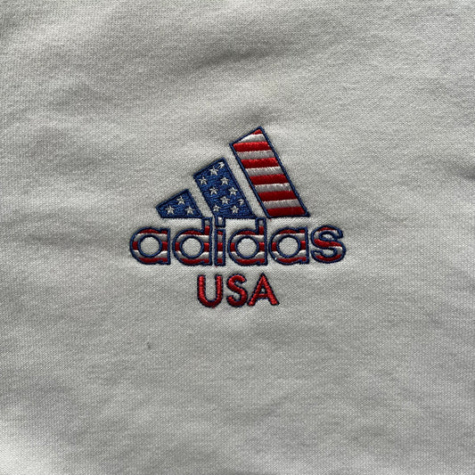 Adidas USA Olympic London 2012 Track Jacket in white with a blue sleeve featuring bronze, silver, and gold adidas stripes. Designed with a red zipper, blue collar, and embroidered adidas USA logo in a flag graphic on the chest, paired with a gold embroidered London 2012 logo on the other side. A rare piece of official Team USA gear from the London Olympic Games.