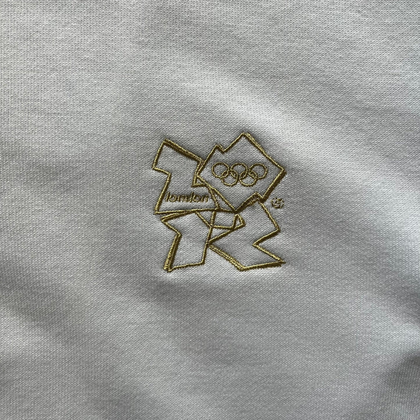 Adidas USA Olympic London 2012 Track Jacket in white with a blue sleeve featuring bronze, silver, and gold adidas stripes. Designed with a red zipper, blue collar, and embroidered adidas USA logo in a flag graphic on the chest, paired with a gold embroidered London 2012 logo on the other side. A rare piece of official Team USA gear from the London Olympic Games.