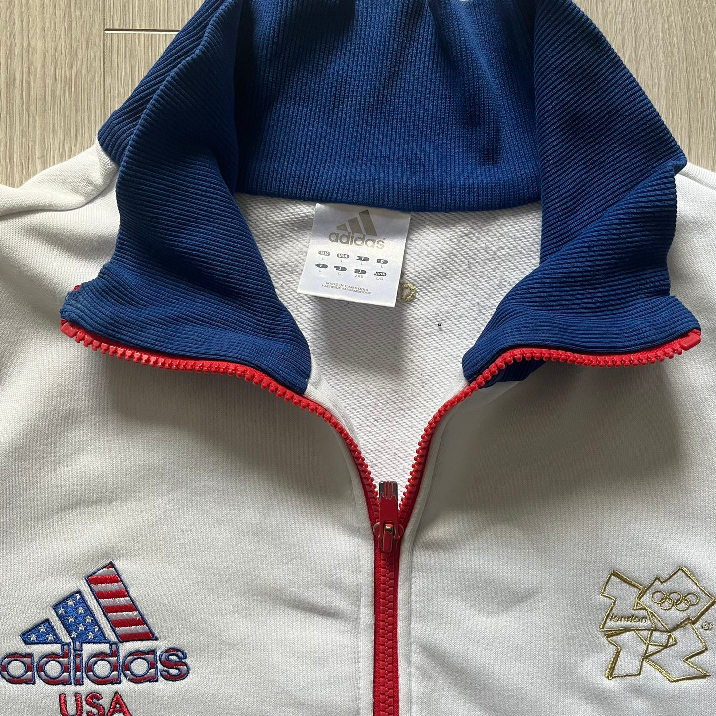 Adidas USA Olympic London 2012 Track Jacket in white with a blue sleeve featuring bronze, silver, and gold adidas stripes. Designed with a red zipper, blue collar, and embroidered adidas USA logo in a flag graphic on the chest, paired with a gold embroidered London 2012 logo on the other side. A rare piece of official Team USA gear from the London Olympic Games.