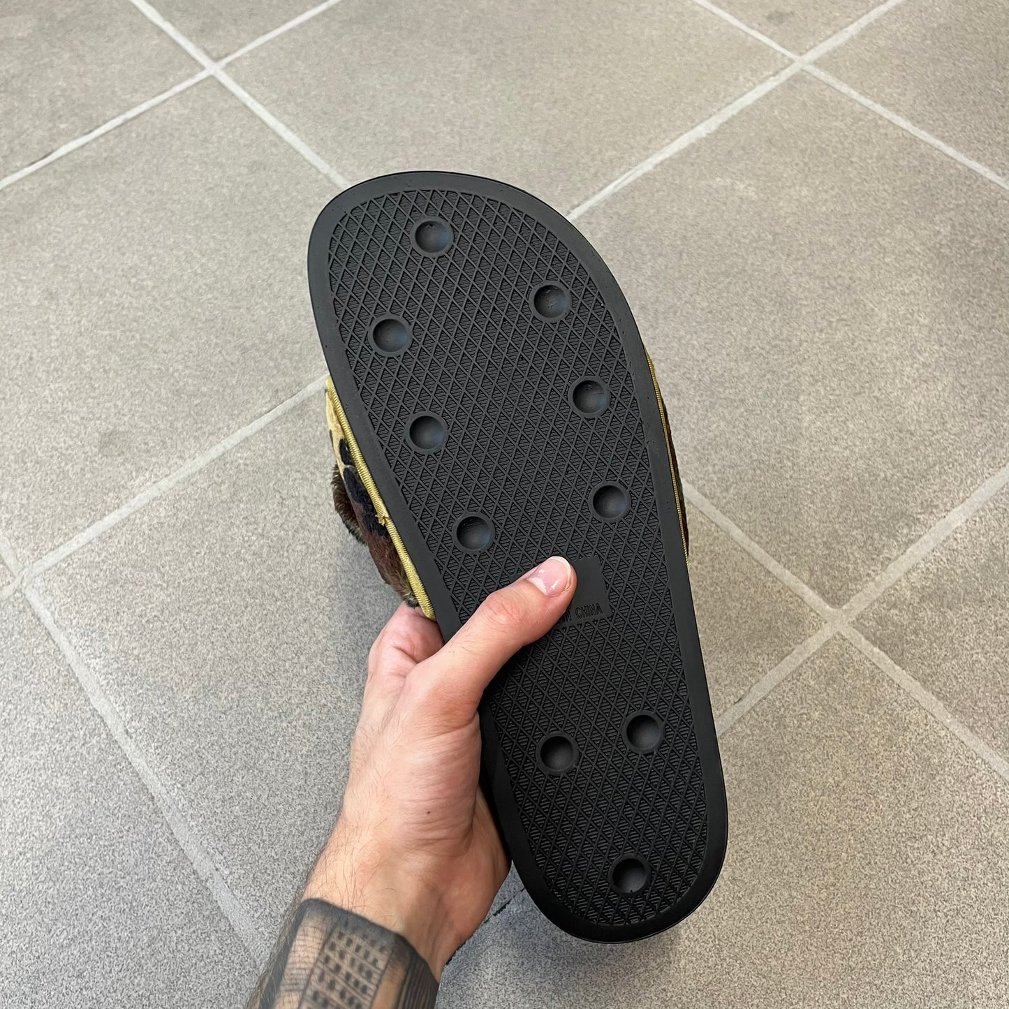 Adidas x Jeremy Scott Adilette Teddy Bear Slides (H02882) were released on November 11, 2021. They feature a black slip-on Adilette silhouette with a camouflage faux fur bandage strap topped by a plush teddy bear head. The strap is lined with leather, while the molded footbed and EVA midsole provide cushioning. Jeremy Scott branding is debossed on the footbed, completing the collaboration design.