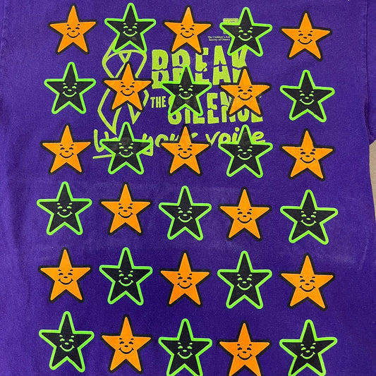 Asspizza 2024 Halloween Stars Purple Tee is a purple T-shirt featuring an all-over star pattern with orange and black stars outlined in green. Each star has a smiling face graphic in the center. The shirt includes a Babbitt 2024 neck tag and is printed on a Children’s Aid blank.