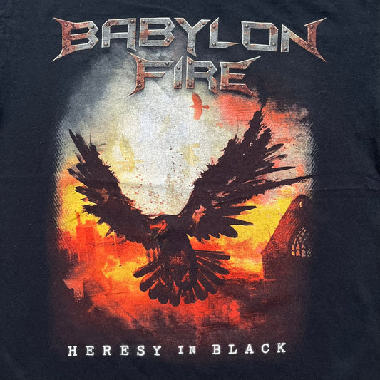 Babylon Fire 2017 “Heresy In Black” Tee is a black T-shirt made on a Gildan blank. The front features the album artwork depicting a fiery landscape with a black bird flying overhead. A stylized metal-inspired “Babylon Fire” logo is printed above the graphic, with “Heresy In Black” text below. Babylon Fire was an alternative rock band active until 2019.