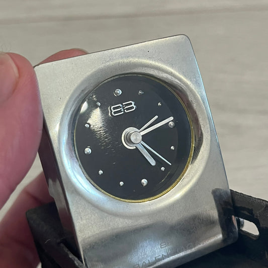 Compact silver-tone metal travel clock with engraved Balenciaga logo. Features a black dial with BB logo and silver-colored hands, plus “Siofor 1000” engraving on the top. Battery-powered and housed in a removable faux leather cover secured by movable side fasteners. Likely produced as a promotional or corporate gift piece.