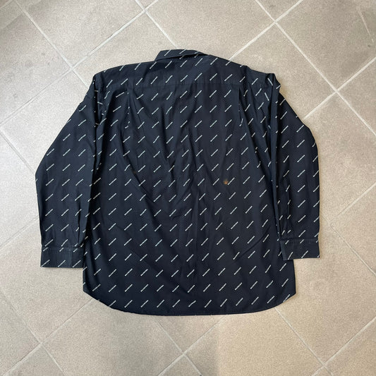 Balenciaga SS18 Black All-Over Logo Shirt is a black button-up made in Italy, featuring a diagonal all-over pattern of small white Balenciaga logo text and black buttons branded with Balenciaga. It has the archetype tag from the Spring/Summer 2018 collection.