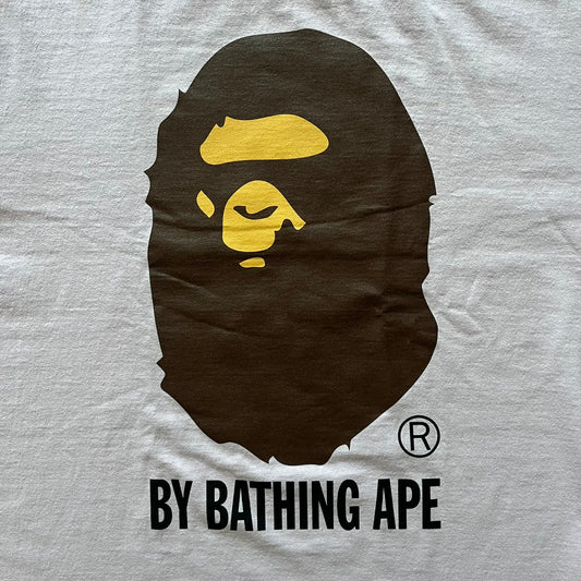 Bape tee in a pinkish/light purple shade, featuring the brand’s classic Ape Head graphic on the front. “By Bathing Ape” text sits underneath for a clean, iconic finish.