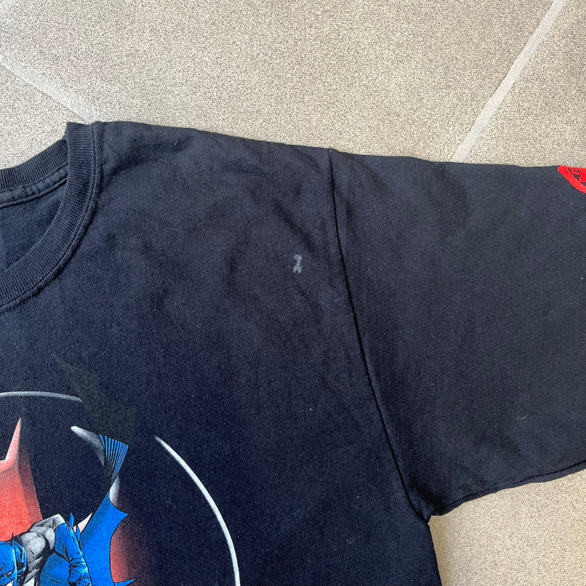 Batman 2005 Graphitti Red Logo Tee is a black T-shirt featuring a graphic of Batman in a blue suit running forward, set against a circular red Bat logo in the background. A red Graphitti logo is placed on one sleeve.