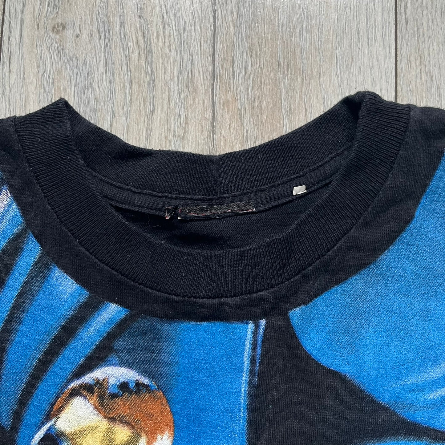 Batman & Robin 1997 AOP Promo Longsleeve Tee is a black long sleeve T-shirt featuring a detailed all-over front print of Batman and Robin in a flying pose, created as part of the official promotional graphics for the film. The design includes a “Batman & Robin” logo at the lower front with a small 1997 licensing print beneath it. The graphic is finished with a thick, paint-like application, giving it a textured, durable feel typical of vintage movie promo apparel.