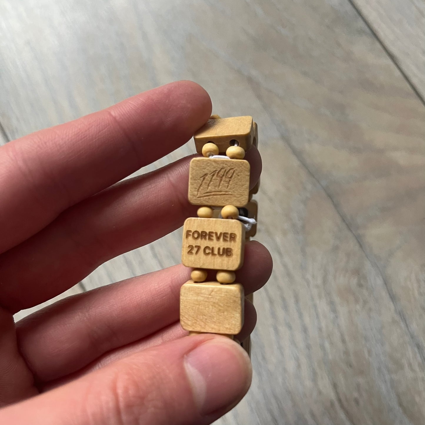 Beige “Forever 27 Club” bracelet by Bazo 1199, showcasing painted wooden beads and engraved portraits of legendary artists from the infamous 27 Club like Kurt Cobain, Basquiat, and Fredo Santana. Signed “forever 27 club” and “1199” on the inside.