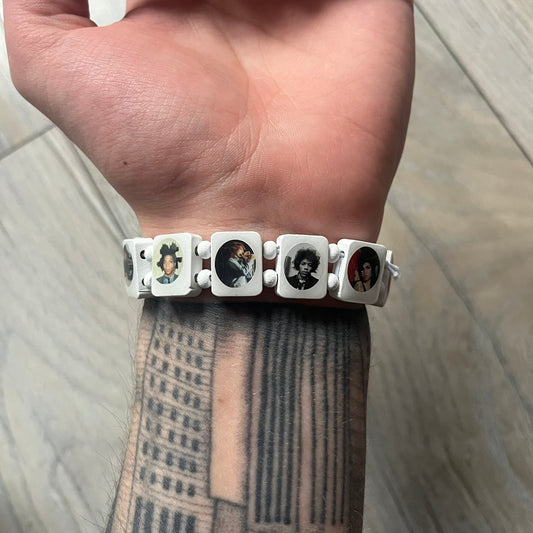 White “Forever 27 Club” bracelet by Bazo 1199, showcasing painted wooden beads and engraved portraits of legendary artists from the infamous 27 Club like Kurt Cobain, Basquiat, and Fredo Santana. Signed “forever 27 club” and “1199” on the inside.
