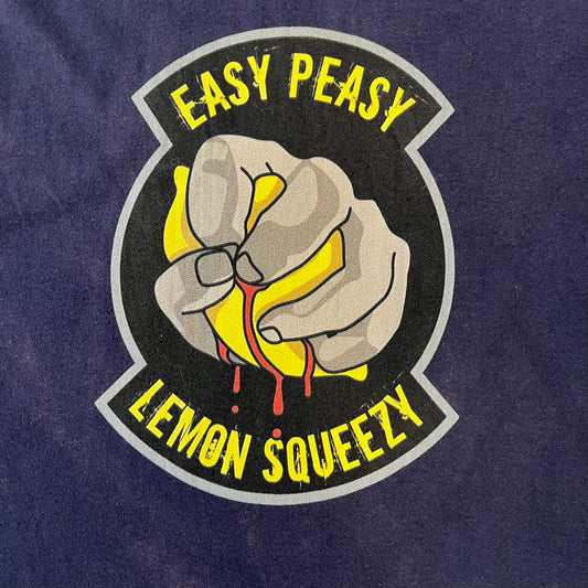 CS:GO Easy Peasy Lemon Squeezy 2015 Tee is a navy T-shirt made in the USA by We Love Fine, featuring a large CS:GO sticker graphic on the front of a hand squeezing a lemon with “Easy Peasy Lemon Squeezy” text. A CS:GO logo is printed on the back below the collar, and it carries a licensed 2015 tag.
