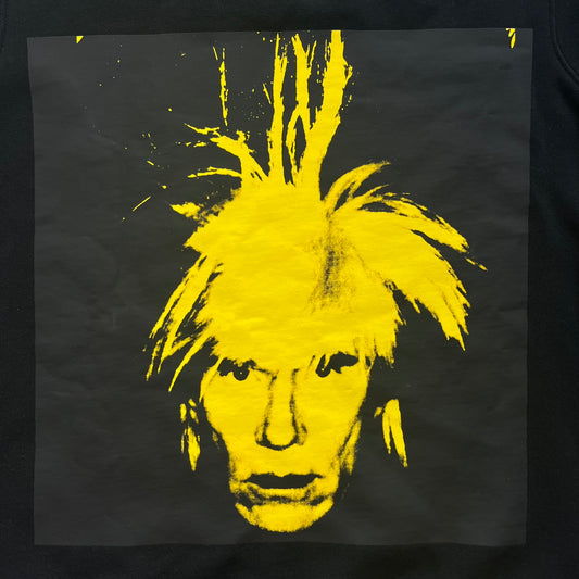 Calvin Klein x Andy Warhol 2018 Self-Portrait Crewneck is a black sweatshirt from the Calvin Klein Jeans x Andy Warhol collaboration. The front features a large yellow self-portrait graphic of Andy Warhol printed on a black background. It includes a yellow Calvin Klein Jeans x Andy Warhol neck tag and a collaboration patch near the bottom hem.