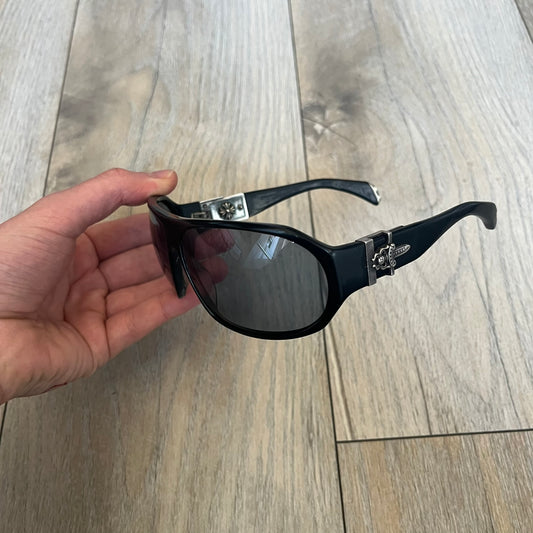 Chrome Hearts Hung Shield Black Sunglasses are black sunglasses from Chrome Hearts featuring black lenses and detailed metal hardware on the arms. The outer arm joints are finished with dagger emblems, while the inner sides feature Chrome Hearts cross motifs. The arm tips include metal end pieces with engraved Chrome Hearts branding. The pair comes with a black leather Chrome Hearts case.