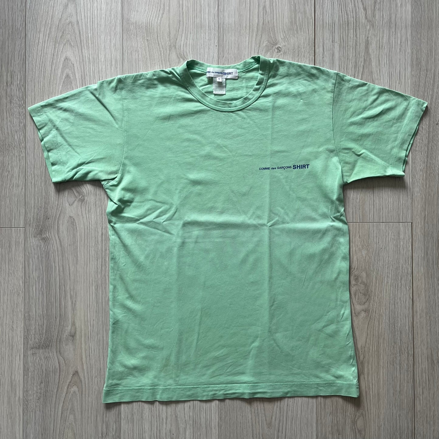 Green t-shirt with a small logo on a wooden floor