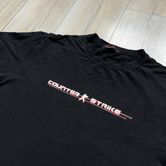 Counter-Strike 2000s Logo Tee is a black T-shirt featuring the classic Counter-Strike logo printed across the front. The design includes white “Counter” and “Strike” text separated by the armed character silhouette, with a red shadow layer behind the logo. A small red Ben Hao tag is attached near the bottom hem.