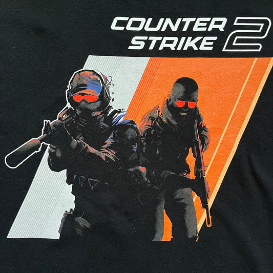 Counter-Strike 2 Promo 2023 Tee is a black T-shirt featuring the game cover graphic on the front, with the Counter-Strike 2 logo above two characters (terrorist and counter-terrorist) set against a white and orange background. The shirt includes a printed CS2 neck tag with 2023 Valve licensing.