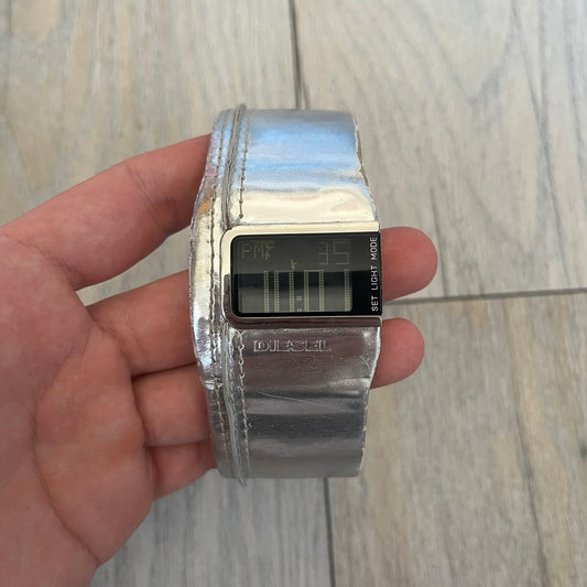 Diesel DZ7090 Silver Leather Strap Digital Watch features a Silver leather patchwork strap and a black digital display housed in a silver rectangular case. The watch includes a backlight, Arabic numeral indices, and a deployant closure, with a stainless steel case back engraved with Diesel branding and model number DZ-7090. It is water resistant up to 50 meters (5 ATM) and combines industrial design with functional quartz digital movement. 