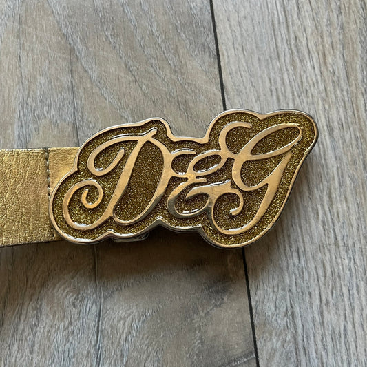 The Dolce & Gabbana 2000’s Gold Tone D&G Logo Belt is made from metallic gold leather and features a large D&G logo buckle shaped directly as the brand’s initials. The buckle uses a cursive D&G logo set over a glittery gold background, framed by a solid gold-tone metal outline. The entire buckle and belt maintain a consistent gold color scheme typical of Dolce & Gabbana accessories from the 2000s.