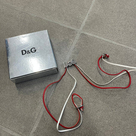 Dolce & Gabbana 2000s Red & White Strings Necklace & Bracelet is a versatile accessory featuring red and white leather cords joined with stainless steel end caps. The center is finished with a stainless steel D&G logo, while the back of the logo features a small engraved D&G mark. Designed to be worn either as a necklace or wrapped as a bracelet, it comes complete with its original box.