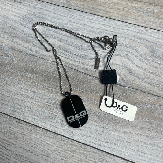 Dolce & Gabbana 2000s Split Black Dog Tag Necklace is a stainless steel necklace featuring a black dog tag pendant with a vertical grey split line running from top to bottom. The lower section of the pendant includes D&G and Dolce & Gabbana logos in the same grey tone as the split detail. The pendant has an iridescent black finish that shifts color depending on the angle. It is attached to a steel ball chain with a lobster clasp closure.