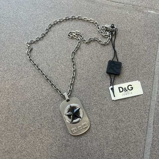 The Dolce & Gabbana Star Dog Tag Necklace (DJ0889) is a heavy stainless steel necklace featuring a thick, oversized dog tag pendant. The front of the pendant has a raised black star mounted on a silver-tone circular base, with open cutout sections between the star and the circle. A raised D&G logo is positioned at the lower front of the dog tag. The necklace is finished with a lobster clasp and has a solid, weighty construction throughout.
