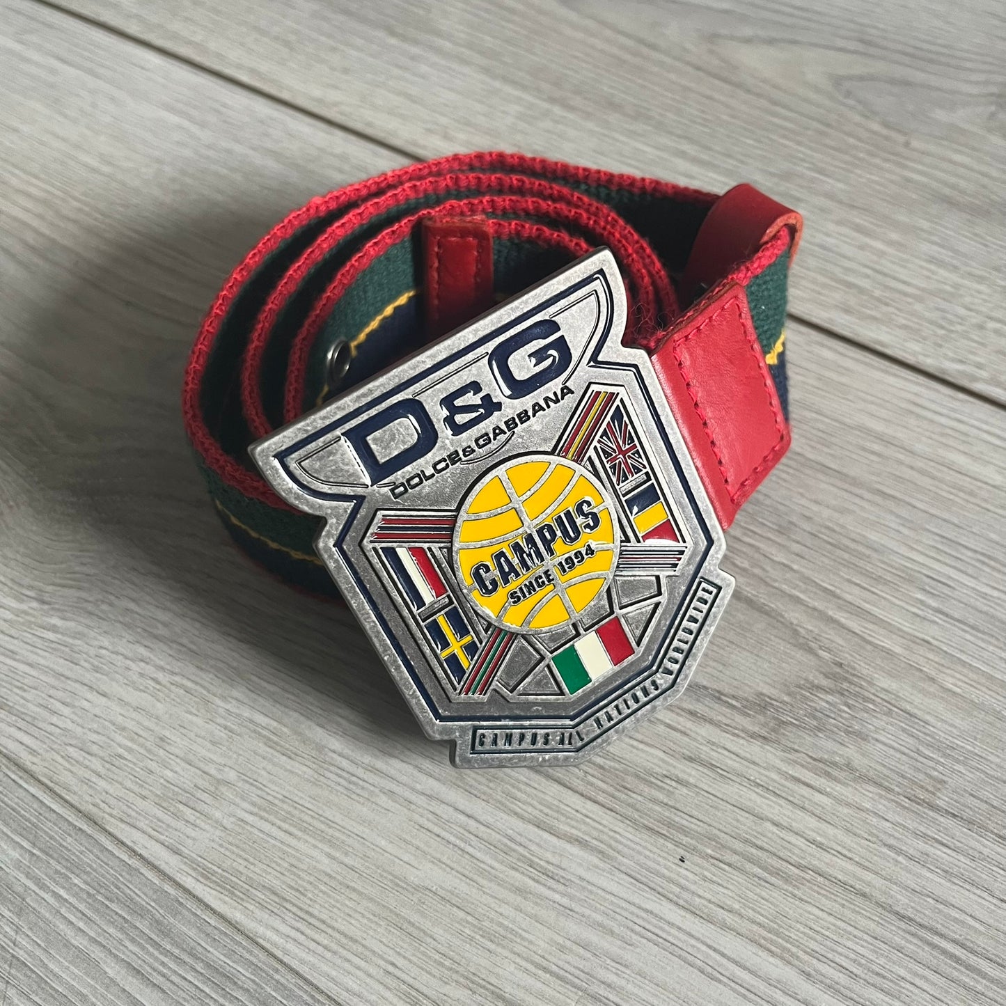 Dolce & Gabbana "Campus Since 1994" Material Belt features a multicolor cotton webbing strap in red, navy, green, and yellow with leather-tipped ends. The statement metal buckle displays a crest design with D&G branding, a central “Campus Since 1994” motif, and multiple national flags. A sporty accessory that combines vintage with luxury detail.