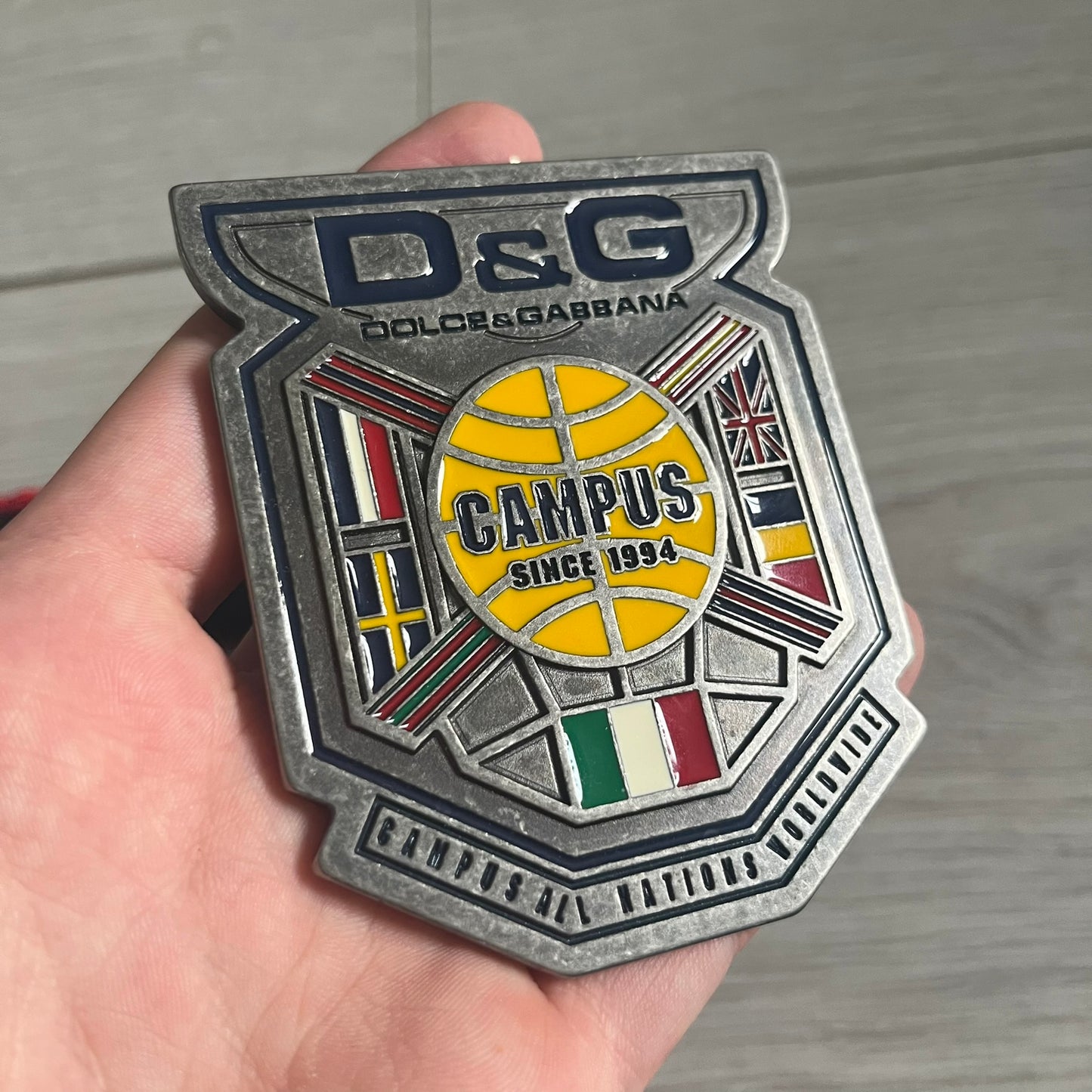 Dolce & Gabbana "Campus Since 1994" Material Belt features a multicolor cotton webbing strap in red, navy, green, and yellow with leather-tipped ends. The statement metal buckle displays a crest design with D&G branding, a central “Campus Since 1994” motif, and multiple national flags. A sporty accessory that combines vintage with luxury detail.
