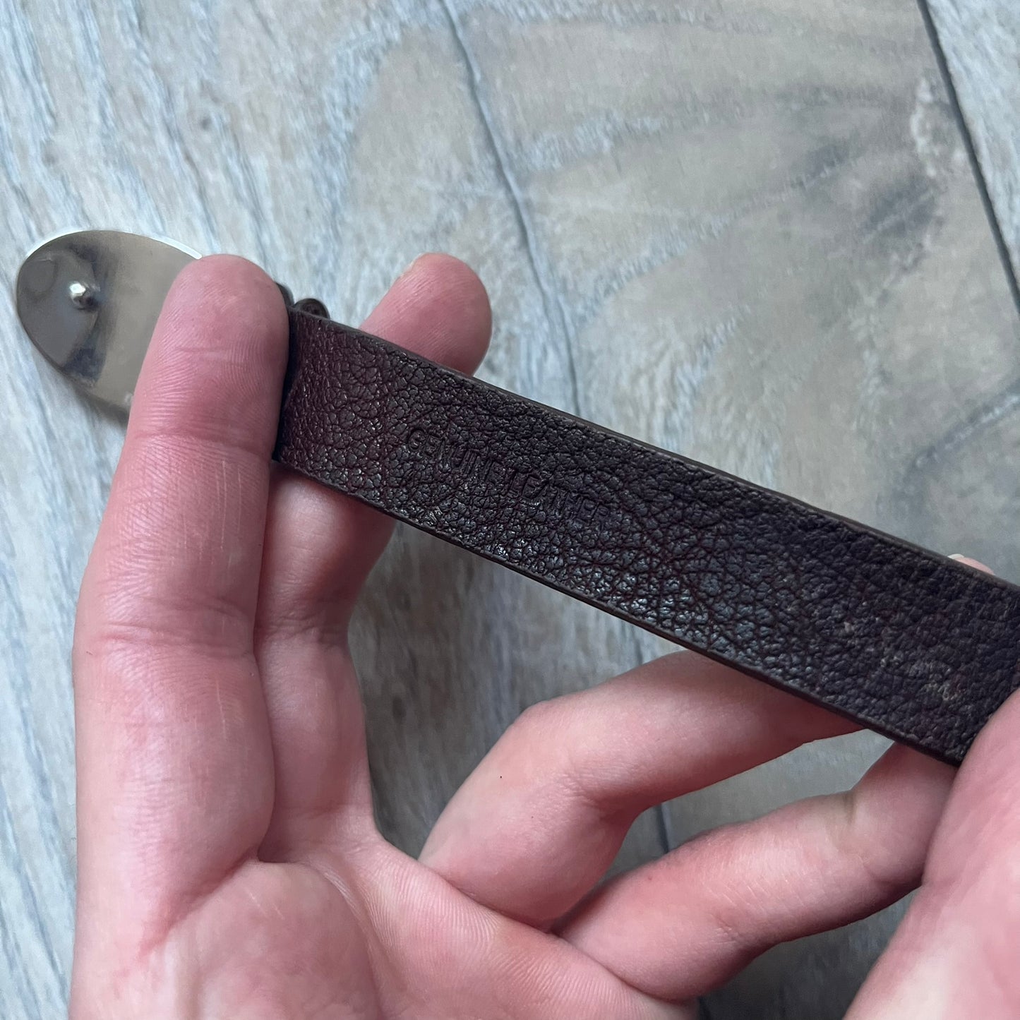 Dolce & Gabbana Champagne Leather Bracelet DJ1097 is a brown leather bracelet designed to resemble a miniature belt, featuring multiple adjustment holes. The silver-tone metal plate is shaped like a D&G champagne label and displays a black D&G logo, along with engraved “Glamorous Champagne 1994 Alc. 12% by Vol.” text. The back of the plate features a large, shallow engraved D&G logo.