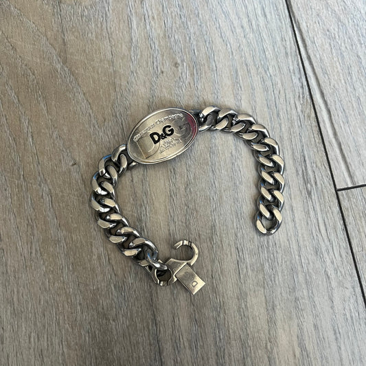 Dolce & Gabbana Champagne Bracelet DJ1092 is a stainless steel bracelet. The silver-tone metal plate is shaped like a D&G champagne label and displays a black D&G logo, along with engraved “Glamorous Champagne 1994 Alc. 12% by Vol.” text. The back of the plate features a large, shallow engraved D&G logo. Retailed at 125 usd in 2000's.