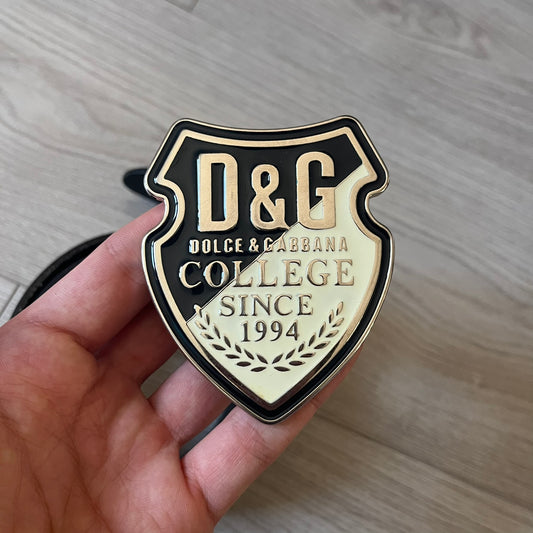 Dolce & Gabbana “College Since 1994” belt featuring a pretty big crest-shaped buckle. The silver text overlays a white-and-black split crest, paired with a soft black leather strap for a refined yet edgy finish.