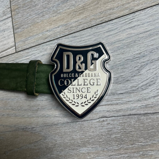 Dolce & Gabbana “College Since 1994” belt featuring a big crest-shaped buckle. The silver text overlays a beige-and-black diagonally split crest, paired with a soft green leather strap in a washed style.