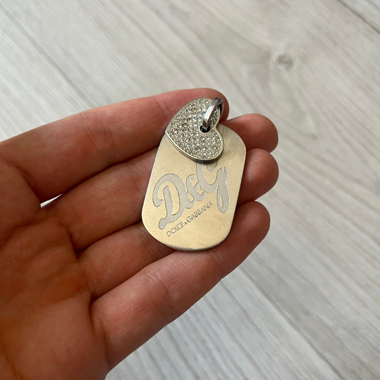 Dolce & Gabbana pendant featuring a silver-tone dog tag engraved with the D&G logo, topped with a separate rhinestone heart charm for a layered, eye-catching effect. Crafted from polished metal, it combines bold branding with a playful, sparkling detail.