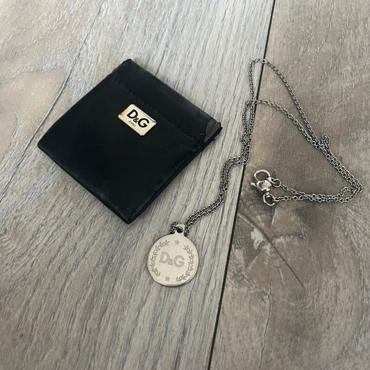Dolce & Gabbana DJ1087 Medal Necklace is a stainless steel chain necklace featuring a circular 2D pendant. The front design includes an engraved D&G logo surrounded by laurel wreath detailing, with small star accents at the top and bottom. The reverse side of the pendant is plain and smooth. It comes with a thin chain, lobster clasp closure, and a black dust bag.
