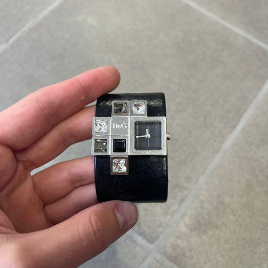 Dolce & Gabbana DW0174 Black Leather Analog Watch features a stainless steel dial composed of multiple squares, including smaller squares set with clear and black crystals, one bearing a D&G logo, and a larger square housing the watch hands. It is water resistant to 3 ATM.
