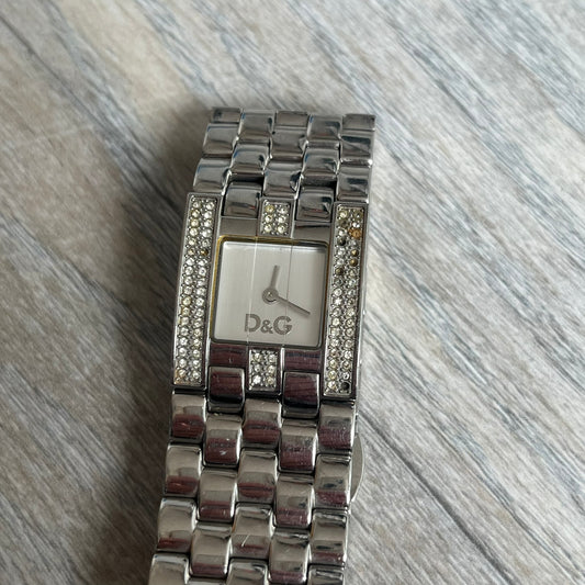Dolce & Gabbana Rhinestones Analog Watch is a stainless steel quartz watch featuring a square white dial with silver D&G logo and silver hands. The case and bracelet form a continuous design with the face matching the width of the bracelet, and selected areas are set with crystal accents. It includes a dual side-clasp closure, is water resistant to 3 ATM, with removable links.