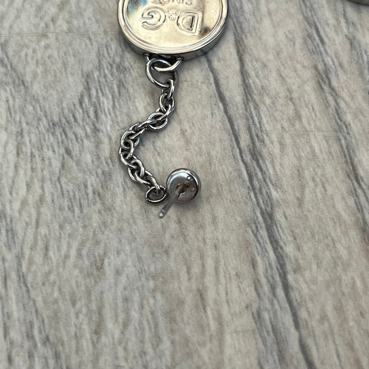 Dolce & Gabbana “D&G Since 1994” Earrings are a pair of 2000s silver-tone hanging earrings. Each earring features a short chain with a round pendant engraved with “D&G Since 1994.” The earrings have a polished metal finish and matching circular plaques.