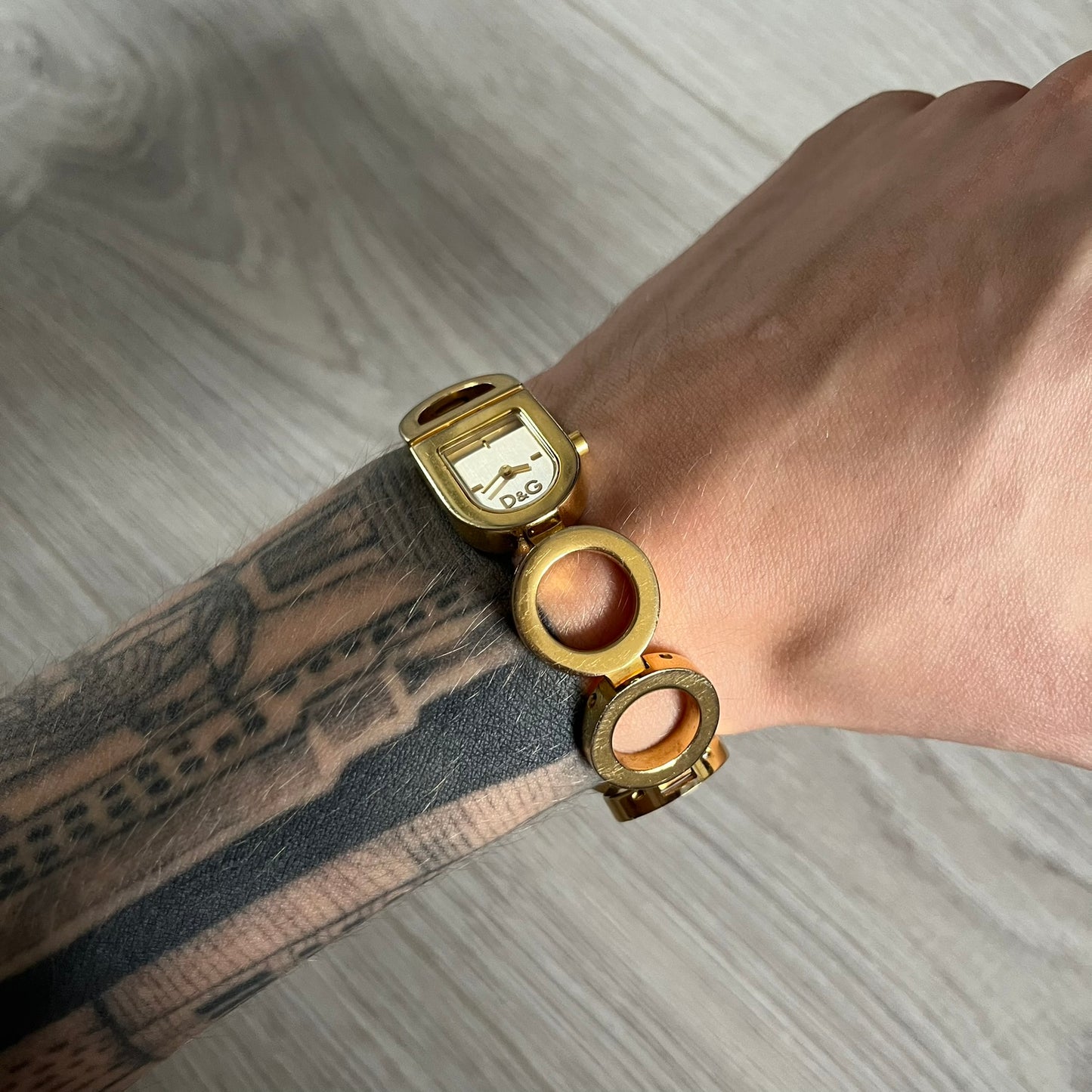 Gold watch and bracelet on a wrist with a tattooed arm, on a light wooden surface.