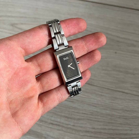 Dolce & Gabbana Holographic Face Analog Watch featuring a slim stainless steel case and a dark face with repeating brown holographic D&G logos. The minimalist dial includes just the small D&G logo and two hands for hours and minutes, giving it a clean and refined look. Water resistant up to 3 ATM, making it both stylish and functional for everyday wear.