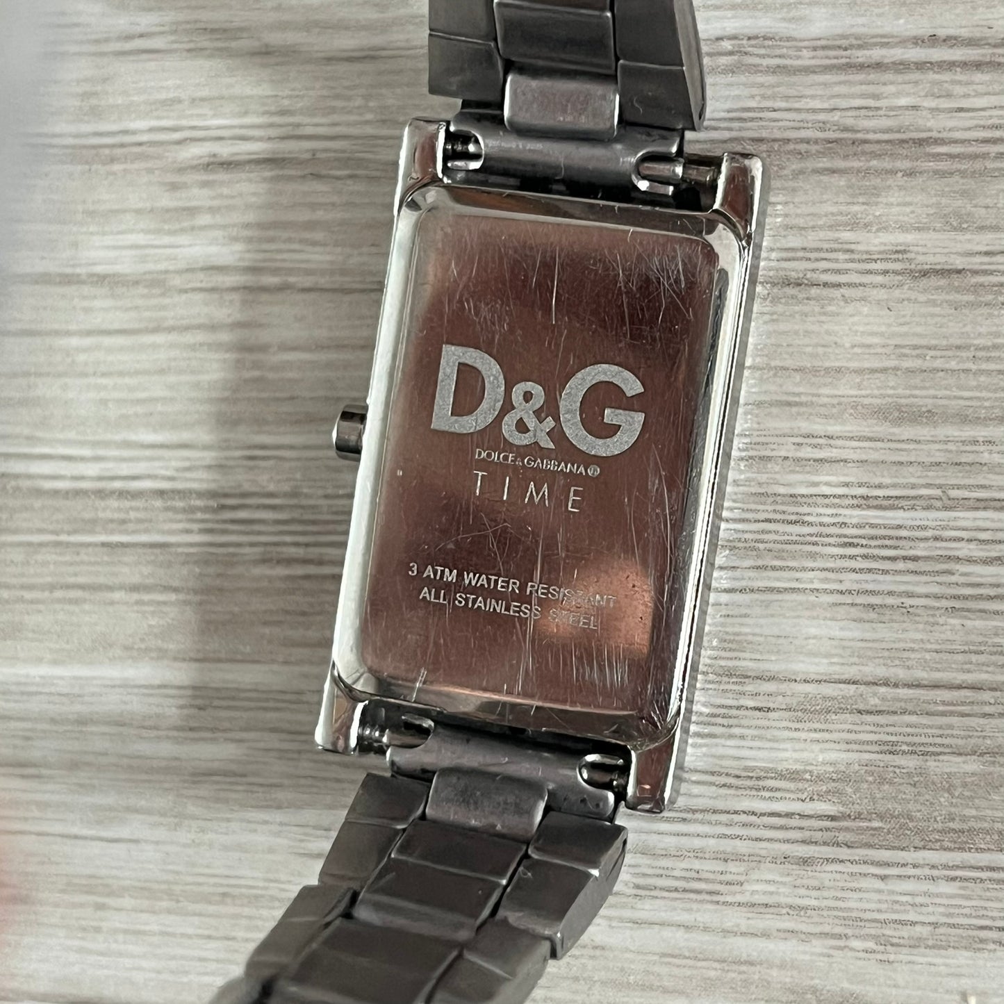 Dolce & Gabbana Holographic Face Analog Watch featuring a slim stainless steel case and a dark face with repeating brown holographic D&G logos. The minimalist dial includes just the small D&G logo and two hands for hours and minutes, giving it a clean and refined look. Water resistant up to 3 ATM, making it both stylish and functional for everyday wear.