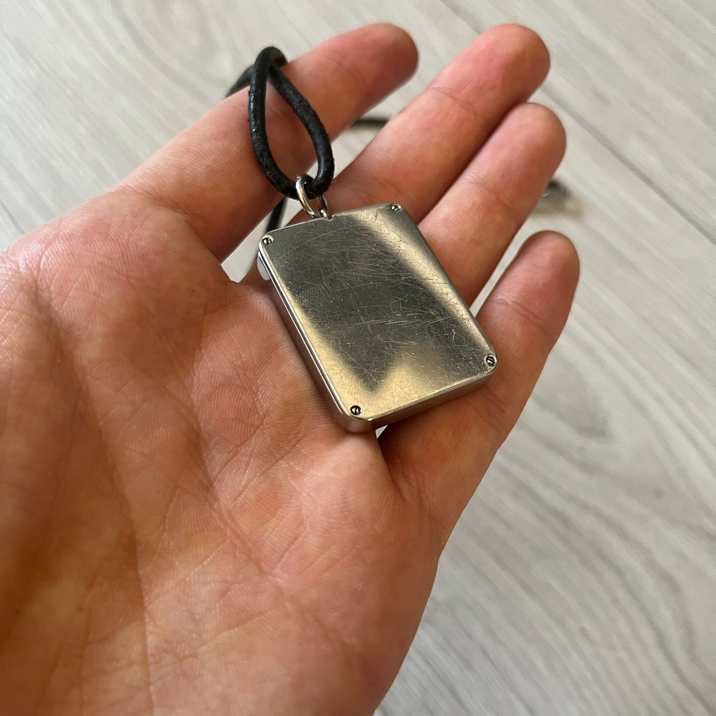 Hand holding a rectangular metallic pendant with a black cord against a neutral background