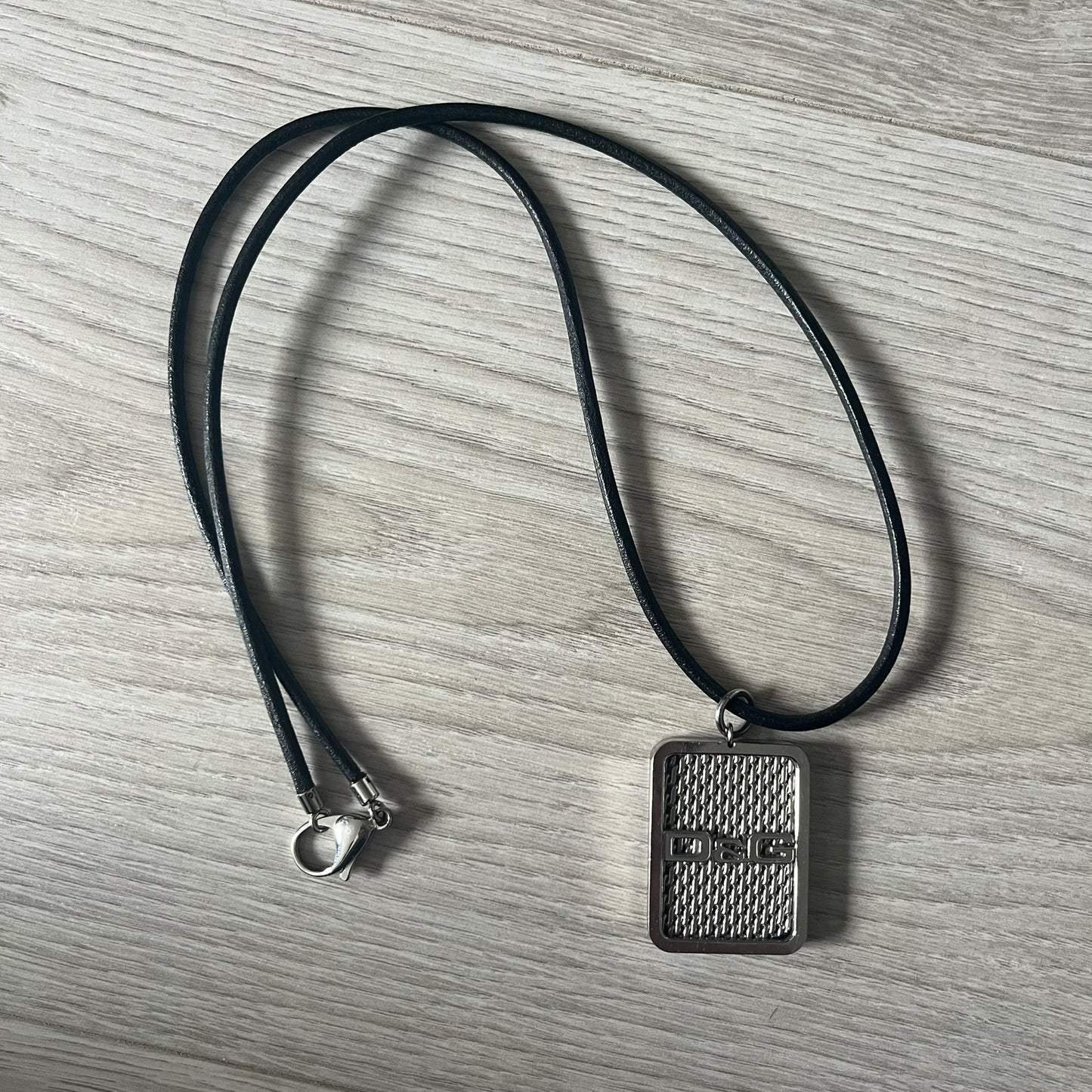 Dolce & Gabbana Mesh Necklace featuring a stainless steel plate engraved with the D&G logo, attached to a black leather cord. The reverse side displays a detailed grid-style mesh design. Model DJ0452, crafted with precision and industrial-inspired elegance.