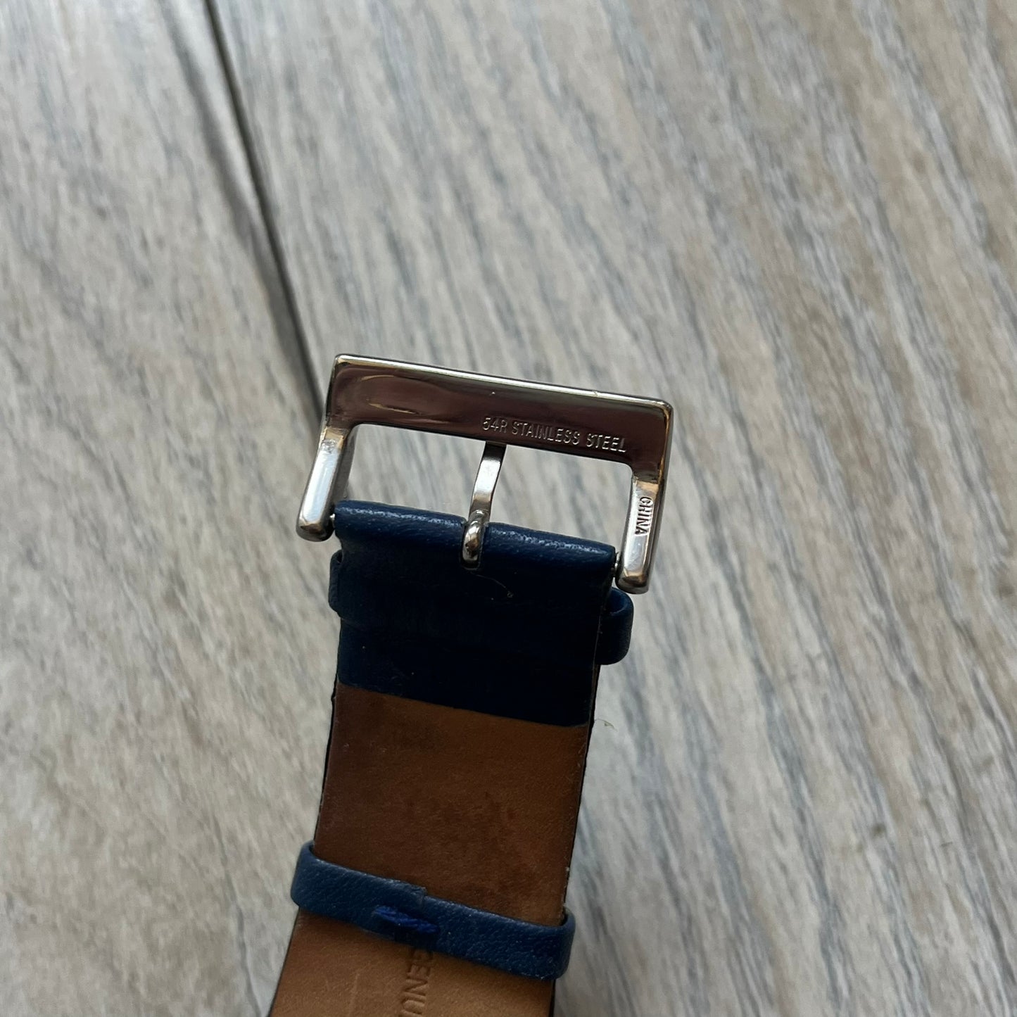 Dolce & Gabbana Navy & Silver Analog Watch features a silver-tone case with a navy dial and matching navy leather strap. The dial has a minimal design with silver hands, a small D&G logo, and a day-date window. The side of the case includes a raised Dolce & Gabbana logo, while the inside of the leather strap is stamped with D&G branding.