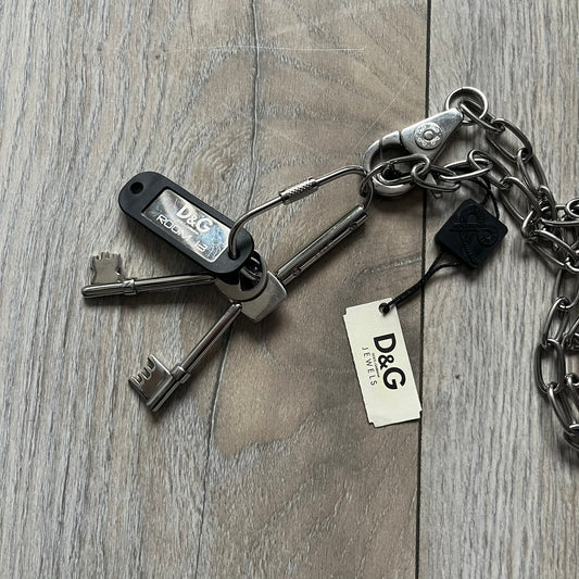 The Dolce & Gabbana Room 13 Carabiner Necklace (DJ0613) is a large, heavy silver-tone necklace featuring a thick paperclip chain with oversized links. At the end of the chain is a large lobster clasp engraved with D&G markings, connected to a branded carabiner. Hanging from the carabiner are two different keys with small D&G logos and a black key identifier with a silver-tone plate engraved with “D&G Dolce & Gabbana Room 13.” The necklace comes with a large silver Dolce & Gabbana dus