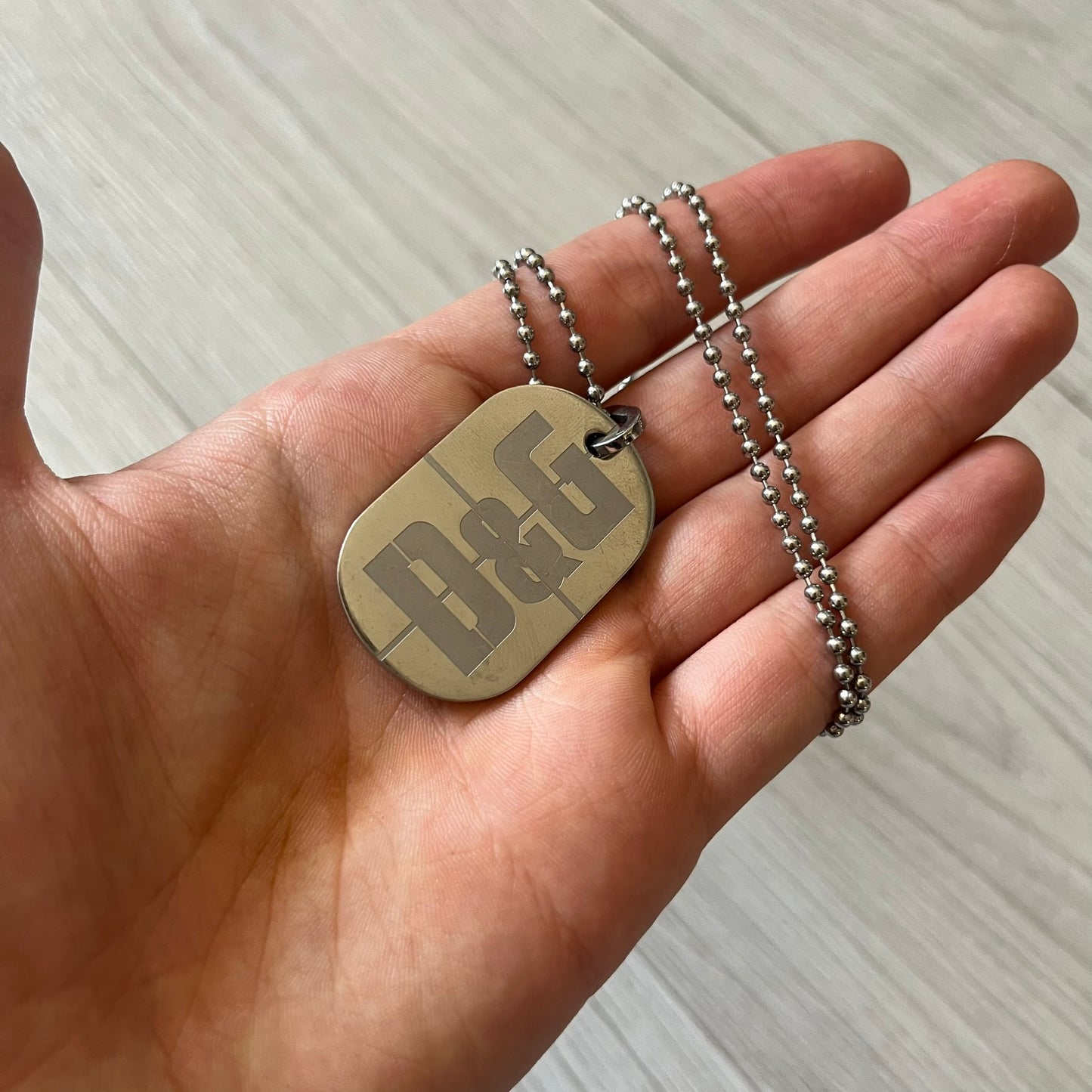 Dolce & Gabbana sniper dog tag necklace in silver-tone stainless steel, featuring a cross resembling a sniper crosshair and a bold D&G logo on top. Completed with a classic ball chain, it merges military-inspired aesthetics with luxury branding. A statement accessory that brings edgy, modern design to any outfit.
