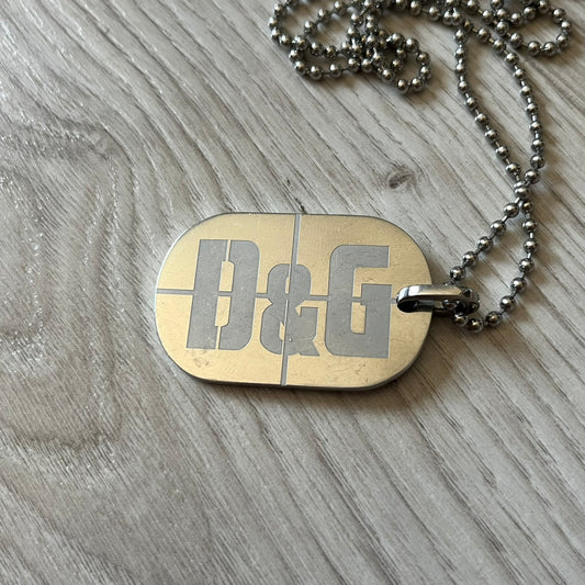 Dolce & Gabbana sniper dog tag necklace in silver-tone stainless steel, featuring a cross resembling a sniper crosshair and a bold D&G logo on top. Completed with a classic ball chain, it merges military-inspired aesthetics with luxury branding. A statement accessory that brings edgy, modern design to any outfit.