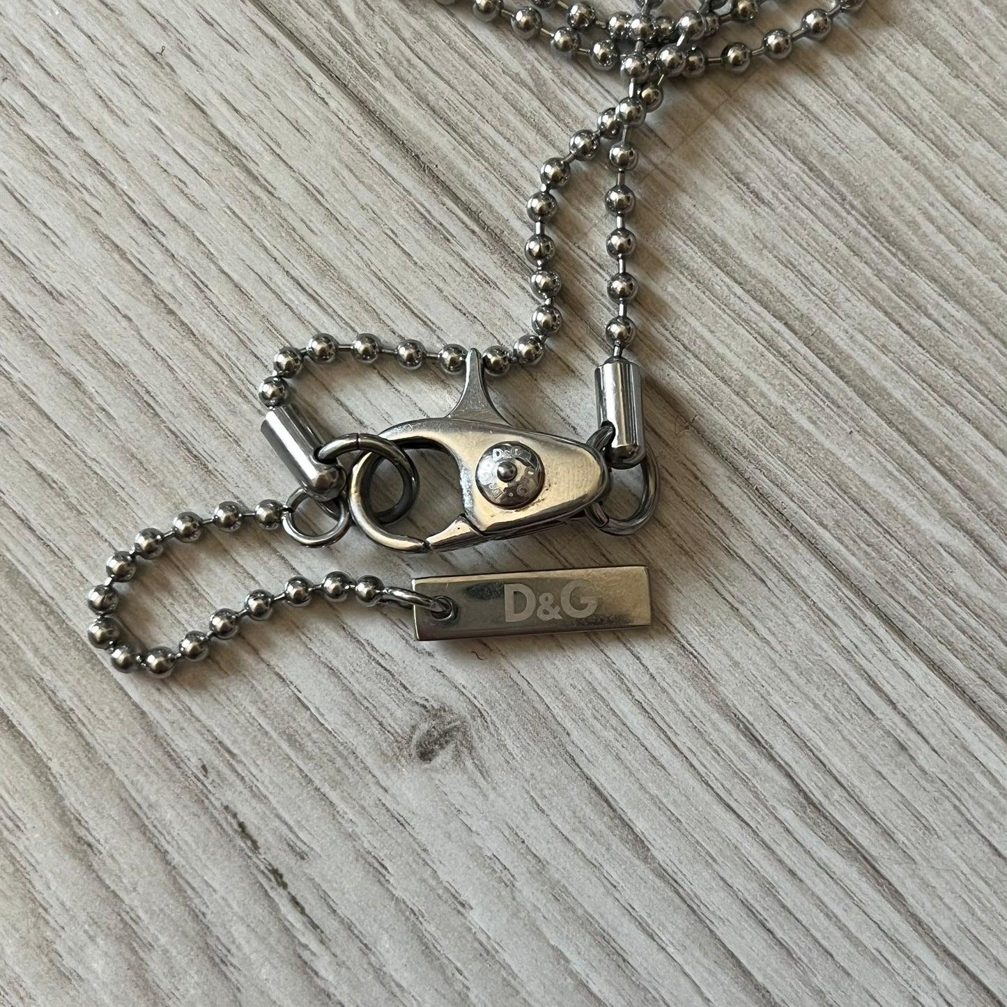 Dolce & Gabbana sniper dog tag necklace in silver-tone stainless steel, featuring a cross resembling a sniper crosshair and a bold D&G logo on top. Completed with a classic ball chain, it merges military-inspired aesthetics with luxury branding. A statement accessory that brings edgy, modern design to any outfit.