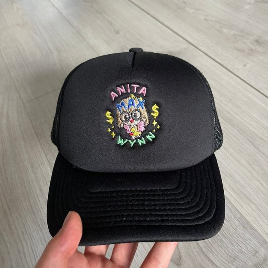 Drake Anita Max Wynn Pink Trucker Hat in all-black front panel, sides and cap, featuring a collection of graphics including a woman with an oversized head, two yellow dollar signs, and “ANITA” in pink, “MAX” in blue, and “WYNN” in green. Designed with a mesh back for breathability, a snapback closure for adjustability, and polyester construction to keep the head cool. A playful tribute to Drake’s alter ego and love of g4mbling.